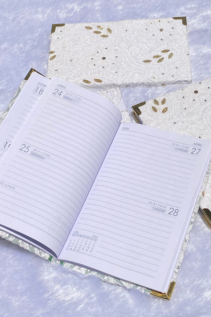 An open white daily planner with lined pages and gold corner accents, styled on a textured light blue surface with matching closed planner covers behind it.