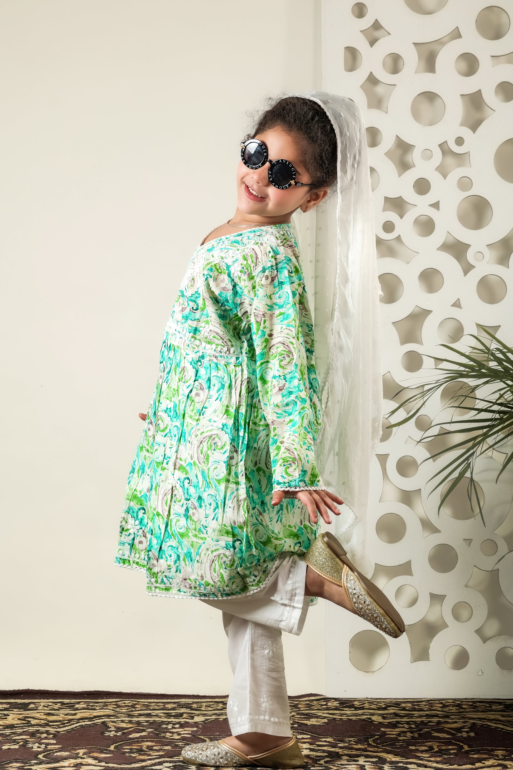 Child wearing a floral outfit with sunglasses and a white scarf against a decorative wall.