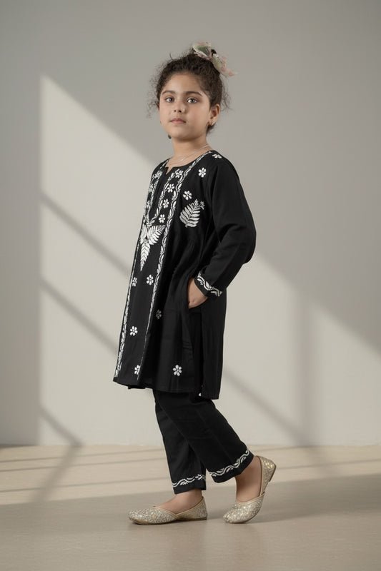 A young girl modeling a black embroidered kurta and matching pants set with white floral and leaf patterns, styled with glittery silver flats against a minimalist beige studio backdrop with soft window shadows.