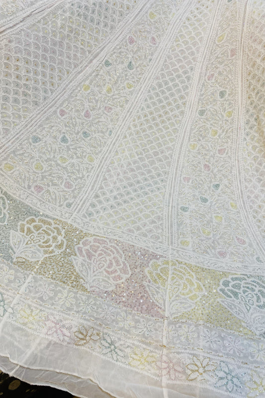 A close-up of a white embroidered fabric, likely a traditional skirt or dupatta, featuring intricate geometric and floral lace patterns with a decorative border.
