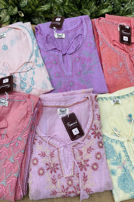 Six folded women's embroidered tops in pastel shades are arranged in two rows on a burlap surface with green foliage in the background. The tops include light pink, lavender purple, coral pink, dusty pink, pale lavender, and pale yellow, all with floral embroidery and tie-neck details.