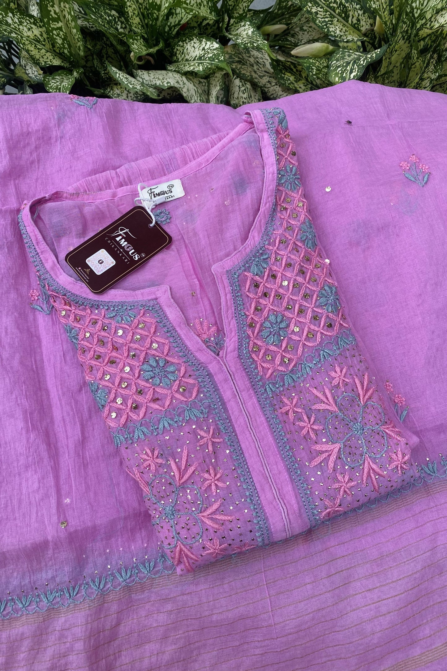A folded lavender purple ethnic kurta set with intricate pink and teal embroidery, displayed on a matching purple dupatta with green foliage in the background.
