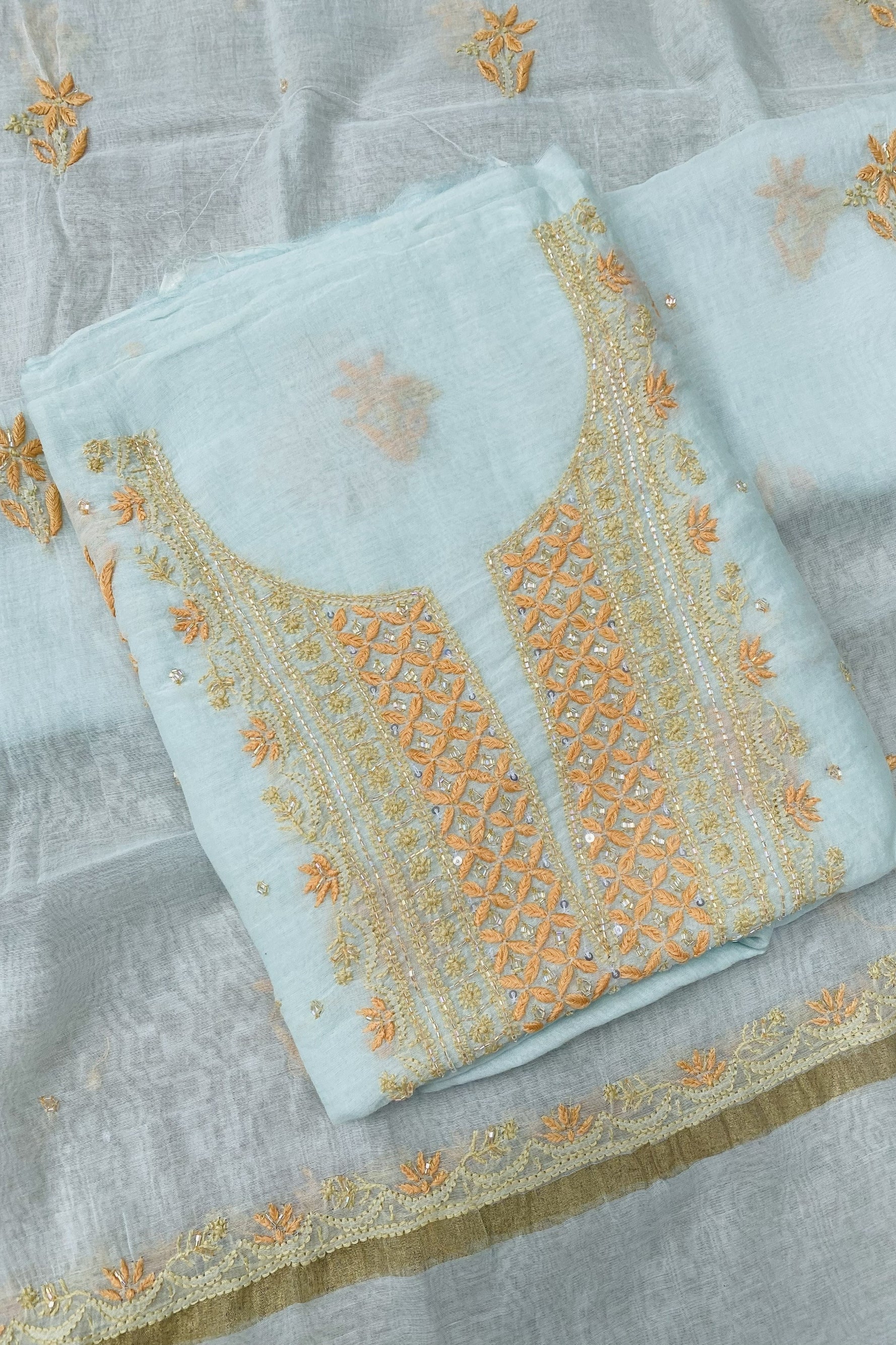 A folded light blue fabric garment with ornate gold and peach embroidery, displayed on a matching sheer fabric background with embroidered details.