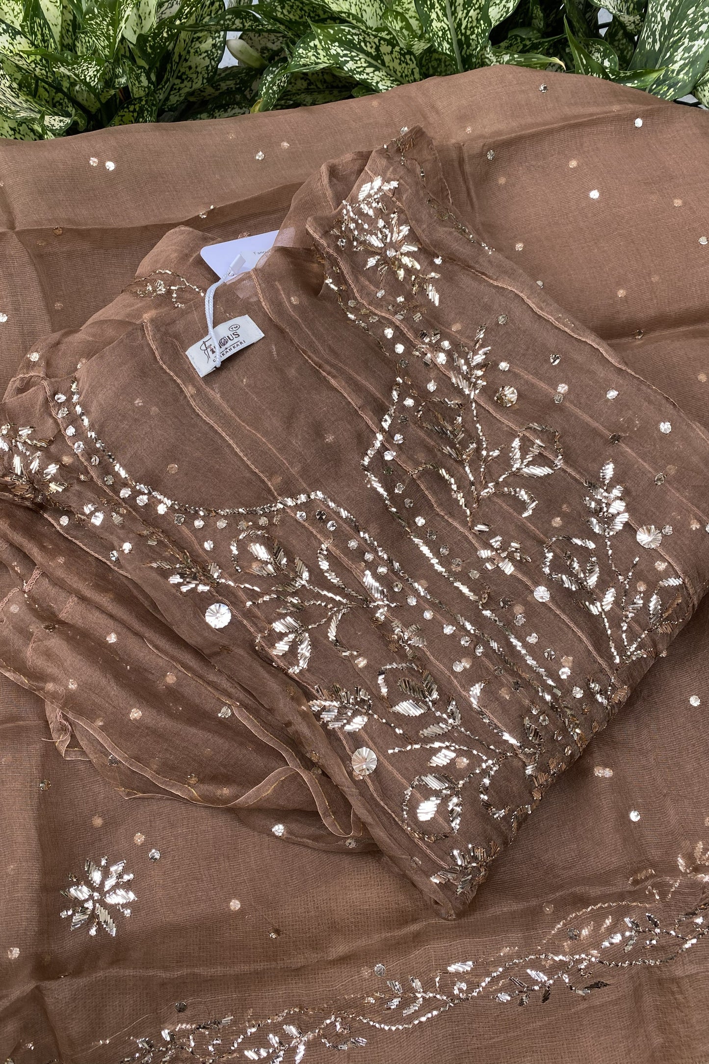 A folded brown organza fabric set with intricate silver mirror work embroidery and sequins, displayed on a matching sheer brown dupatta with scattered silver embellishments.