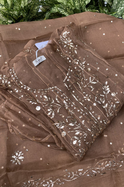 A folded brown organza fabric set with intricate silver mirror work embroidery and sequins, displayed on a matching sheer brown dupatta with scattered silver embellishments.