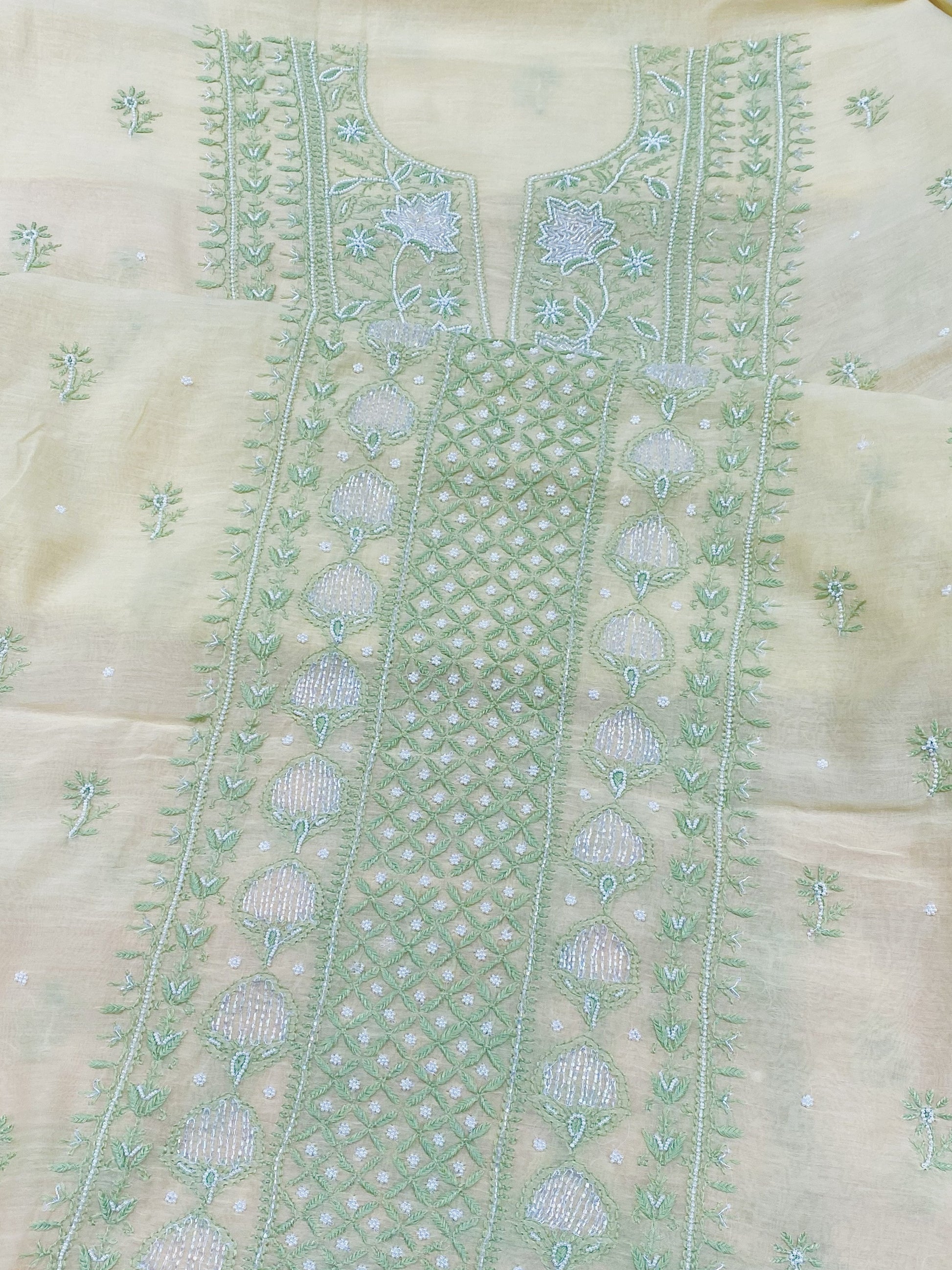 A close-up of a light beige or cream-colored embroidered fabric, likely part of an unstitched salwar suit set. The fabric features a prominent vertical panel with intricate mint green and light blue floral embroidery, geometric patterns, and small scattered motifs.
