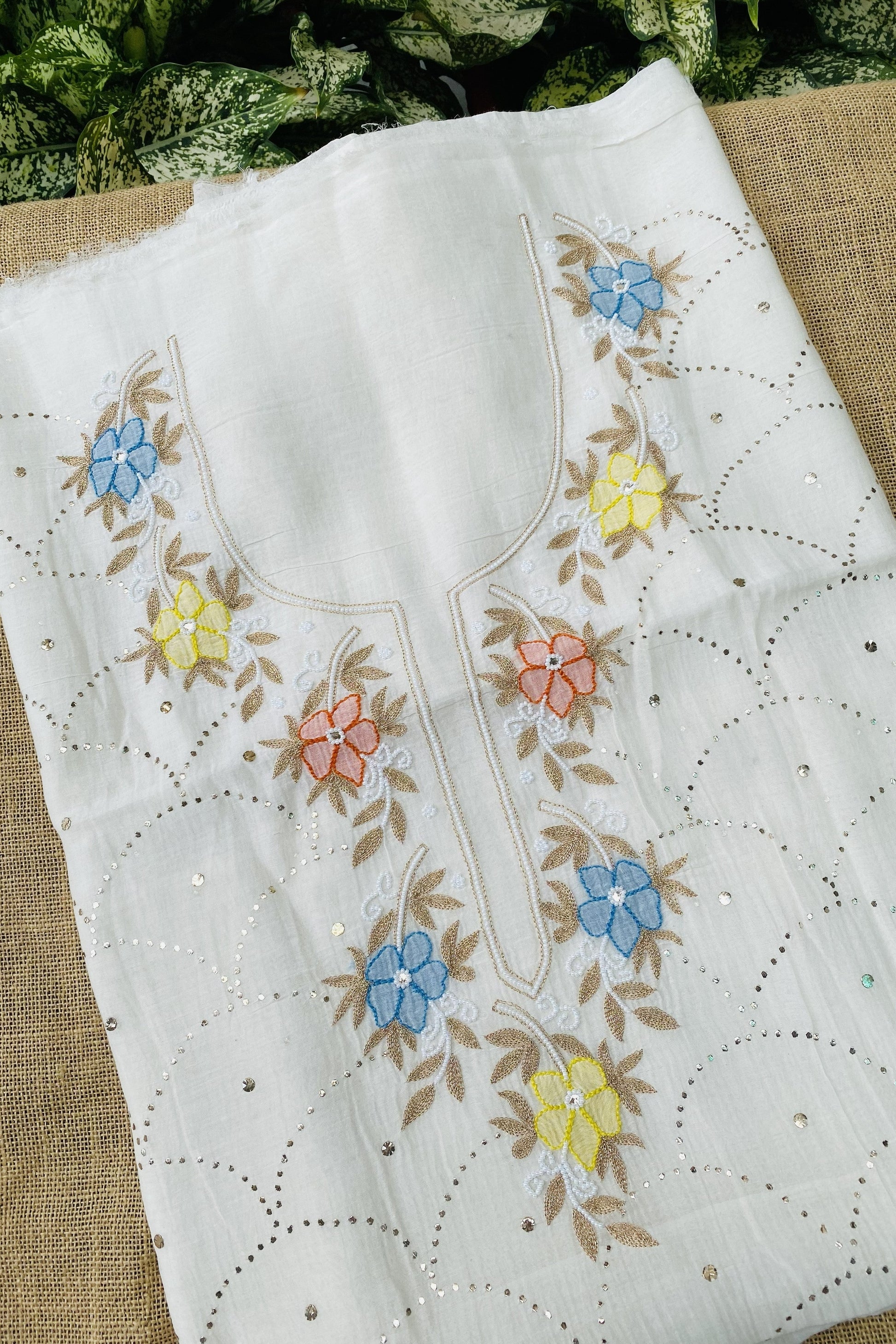 White cotton fabric with colorful floral embroidery in blue, yellow, and pink, featuring scalloped eyelet borders and small silver mirror embellishments, laid flat on a burlap surface.
