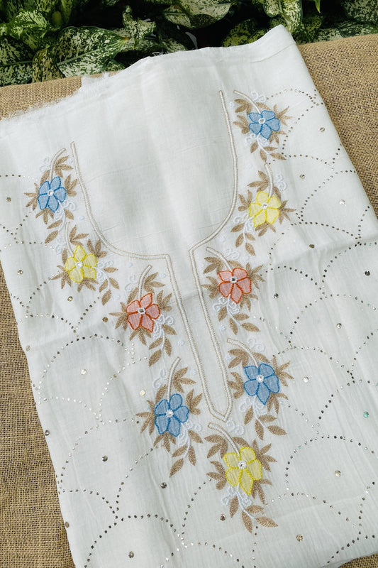 White cotton fabric with colorful floral embroidery in blue, yellow, and pink, featuring scalloped eyelet borders and small silver mirror embellishments, laid flat on a burlap surface.