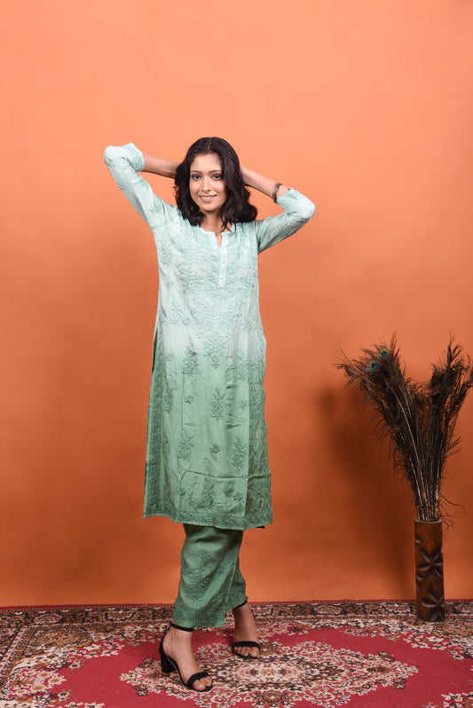 Woman wearing a green traditional outfit against an orange wall with a decorative rug and plant.