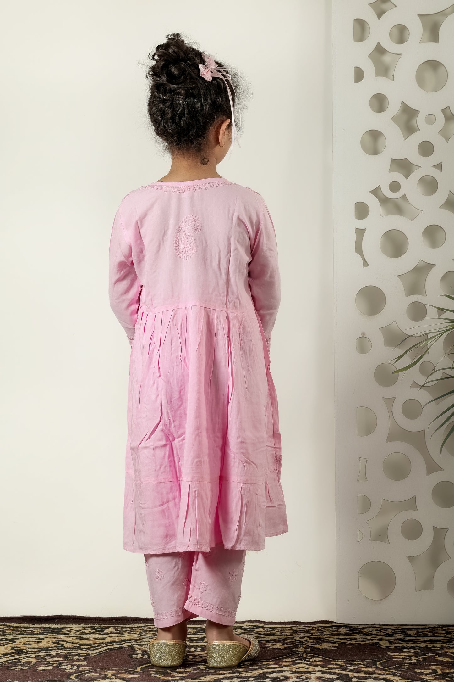 Child wearing a pink traditional outfit standing against a white wall with decorative patterns.