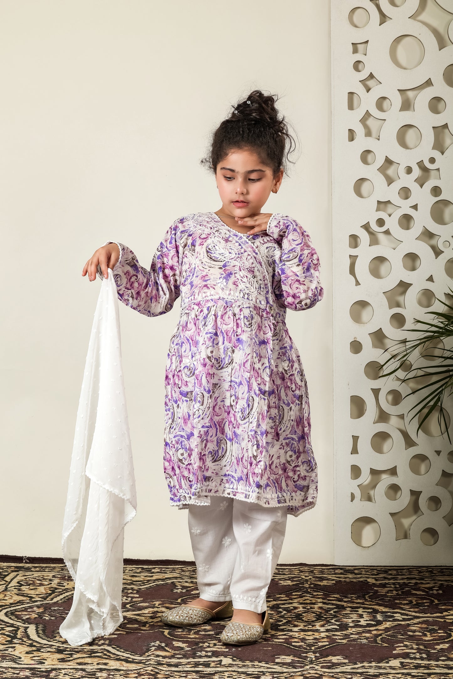 Young girl in a patterned kurta with white pants standing against a decorative wall.