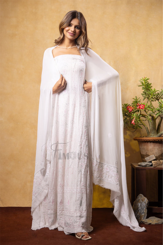 Woman in a white dress holding a white shawl against a beige wall with a plant in the background.