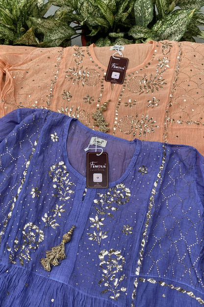 Two women's embroidered tunic tops displayed together, with one in dusty blue in the foreground and one in peach-pink in the background. Both feature intricate silver mirror-work embroidery, sequins, and tassels.