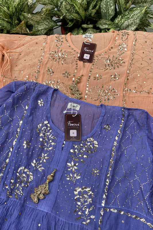 Two women's embroidered tunic tops displayed together, with one in dusty blue in the foreground and one in peach-pink in the background. Both feature intricate silver mirror-work embroidery, sequins, and tassels.