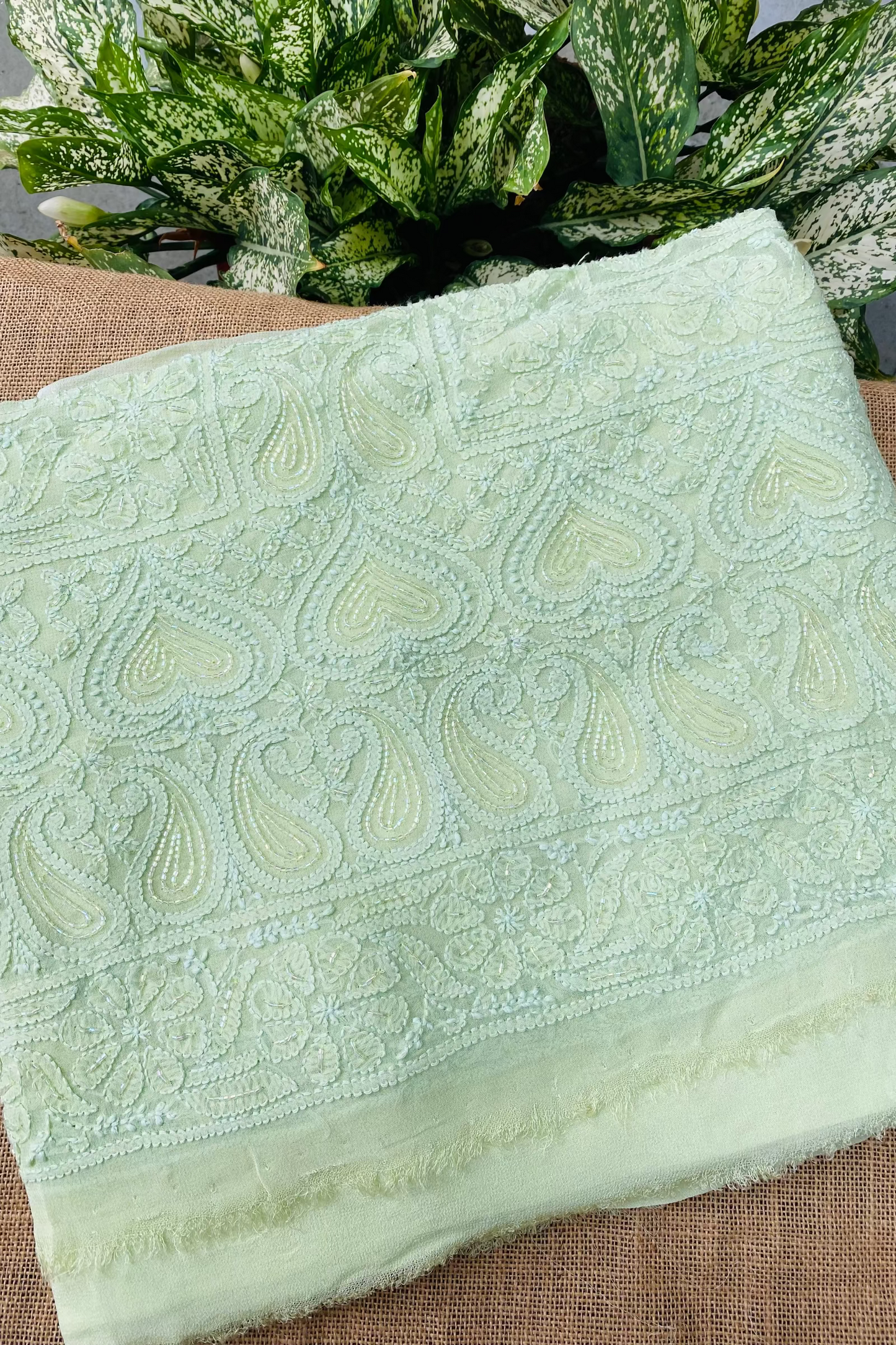 A folded light green embroidered fabric piece with detailed paisley and floral stitching, displayed on a brown woven surface with green leafy plants in the background.