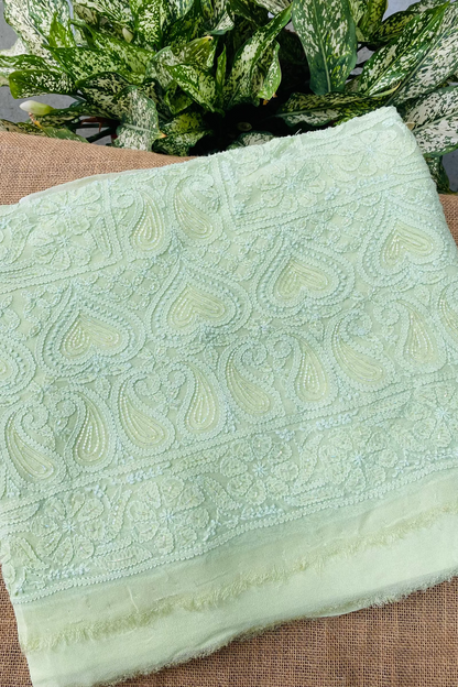 A folded light green embroidered fabric piece with detailed paisley and floral stitching, displayed on a brown woven surface with green leafy plants in the background.
