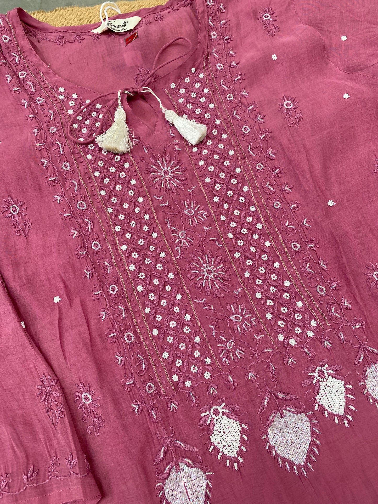 A dusty pink embroidered kurta laid flat, featuring intricate white and silver thread embroidery, tassel ties at the neckline, and heart-shaped embroidered motifs near the hem.