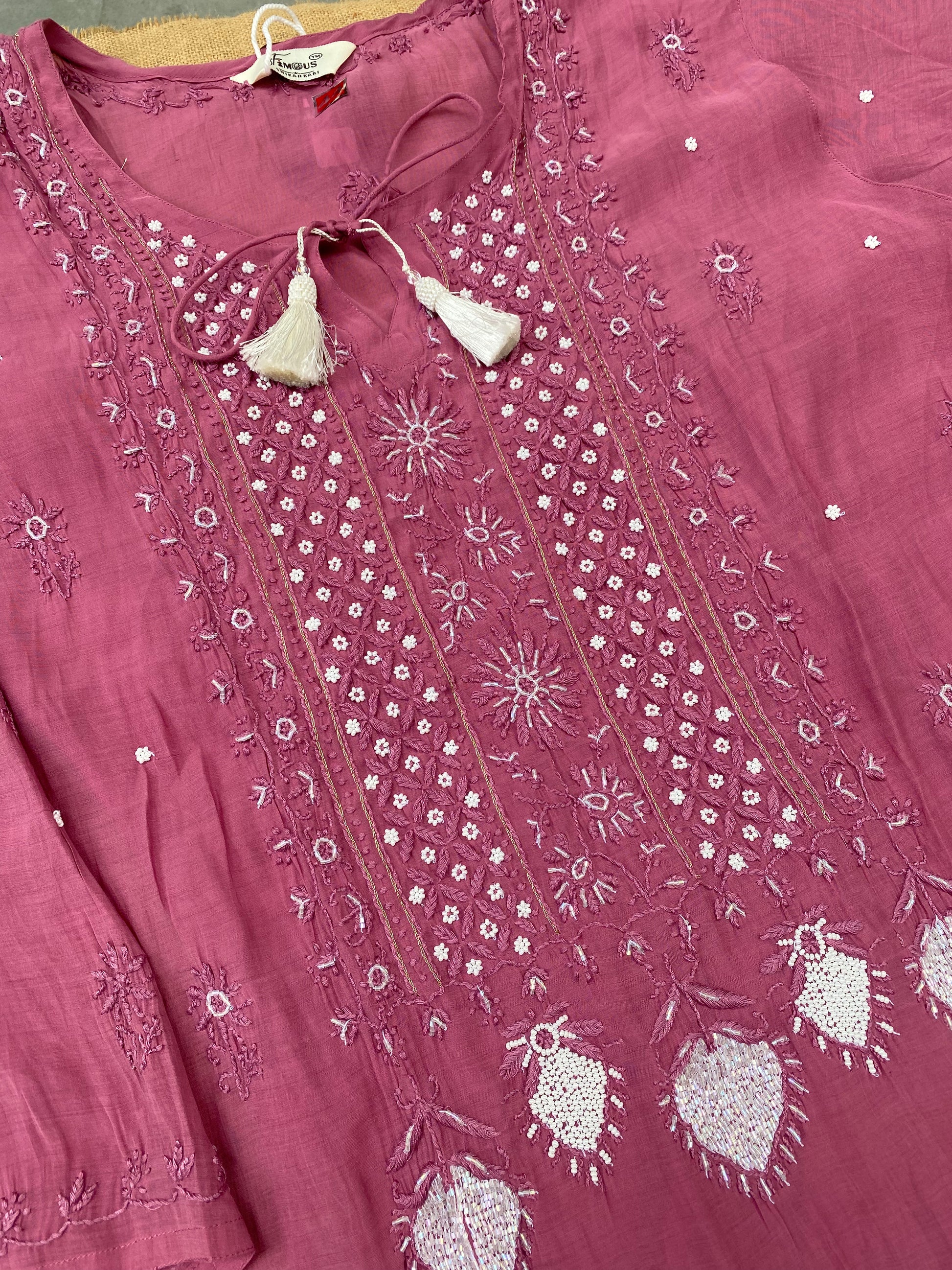 A dusty pink embroidered kurta laid flat, featuring intricate white and silver thread embroidery, tassel ties at the neckline, and heart-shaped embroidered motifs near the hem.