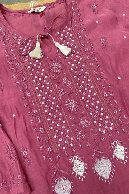 A dusty pink embroidered kurta laid flat, featuring intricate white and silver thread embroidery, tassel ties at the neckline, and heart-shaped embroidered motifs near the hem.