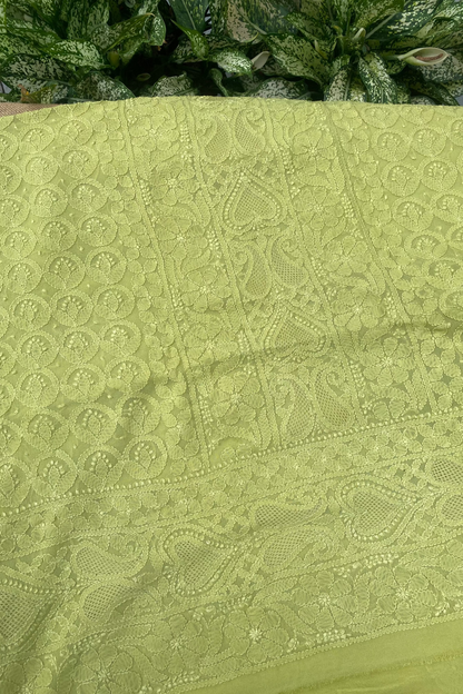 A close-up of a light olive green embroidered fabric with intricate floral and paisley lace patterns, laid flat with green foliage visible in the background.