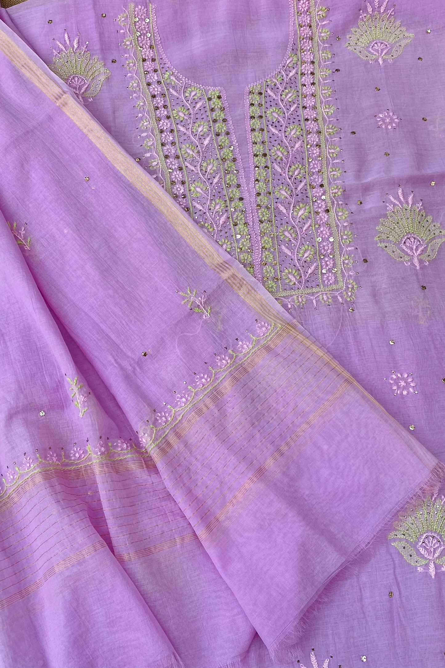 A close-up of a lavender purple unstitched salwar suit fabric set, featuring a kurta with detailed green and gold embroidery on the neckline and a matching dupatta with a patterned border.