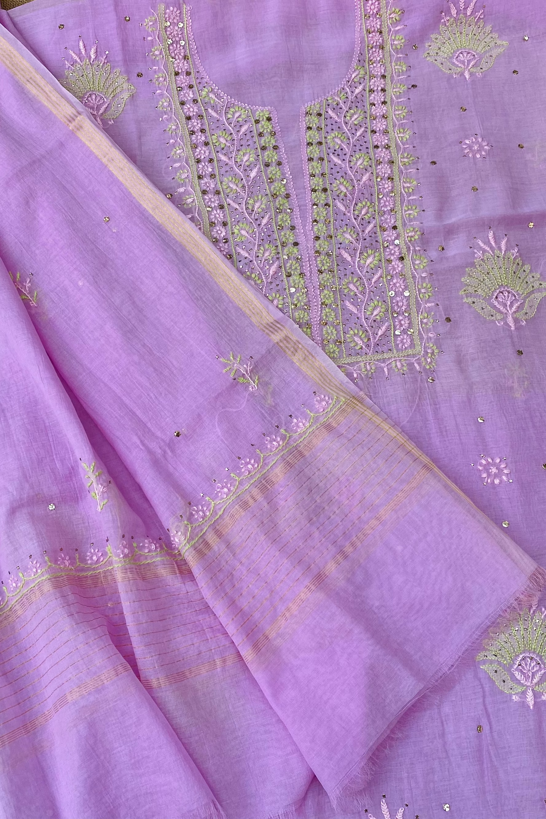 A close-up of a lavender purple unstitched salwar suit fabric set, featuring a kurta with detailed green and gold embroidery on the neckline and a matching dupatta with a patterned border.