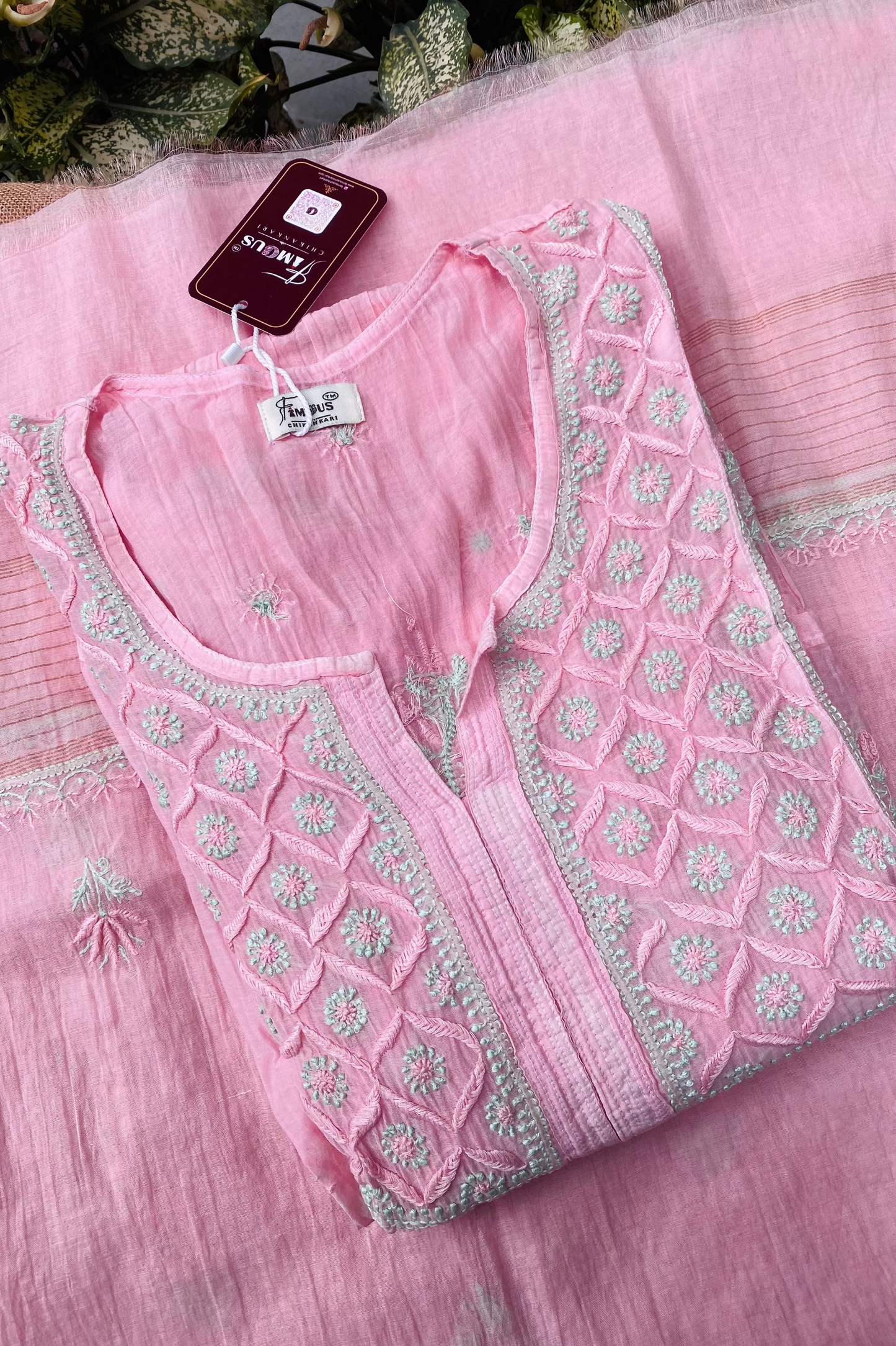 A folded light pink embroidered kurta laid flat on a matching pink fabric background. The kurta has a round neckline, a split V-neckline, and detailed geometric and floral embroidery in pink and light green around the neckline and front panel.
