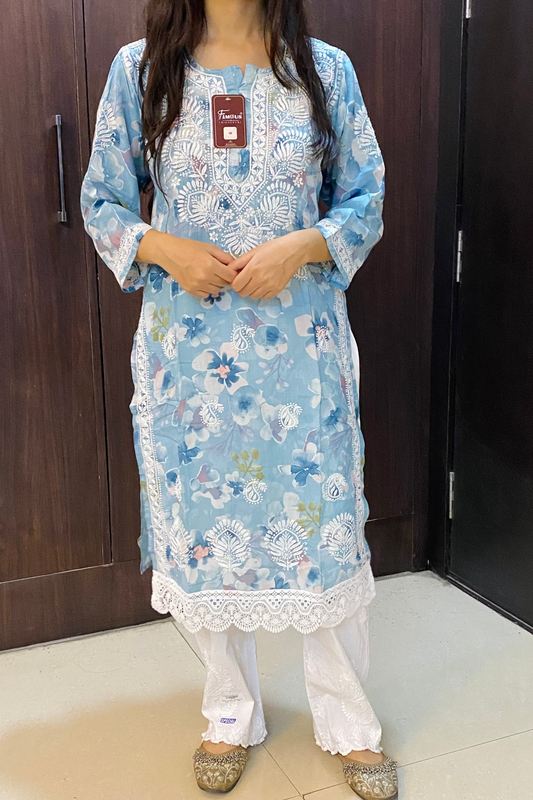 A woman modeling a light blue floral embroidered kurta with white lace trim, paired with white pants and brown shoes, standing in front of dark wooden doors.