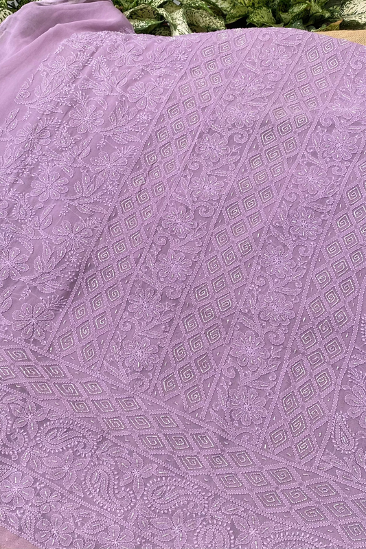 A close-up of a lavender purple embroidered fabric with intricate geometric and floral lace patterns, draped over a surface with green foliage visible in the background.