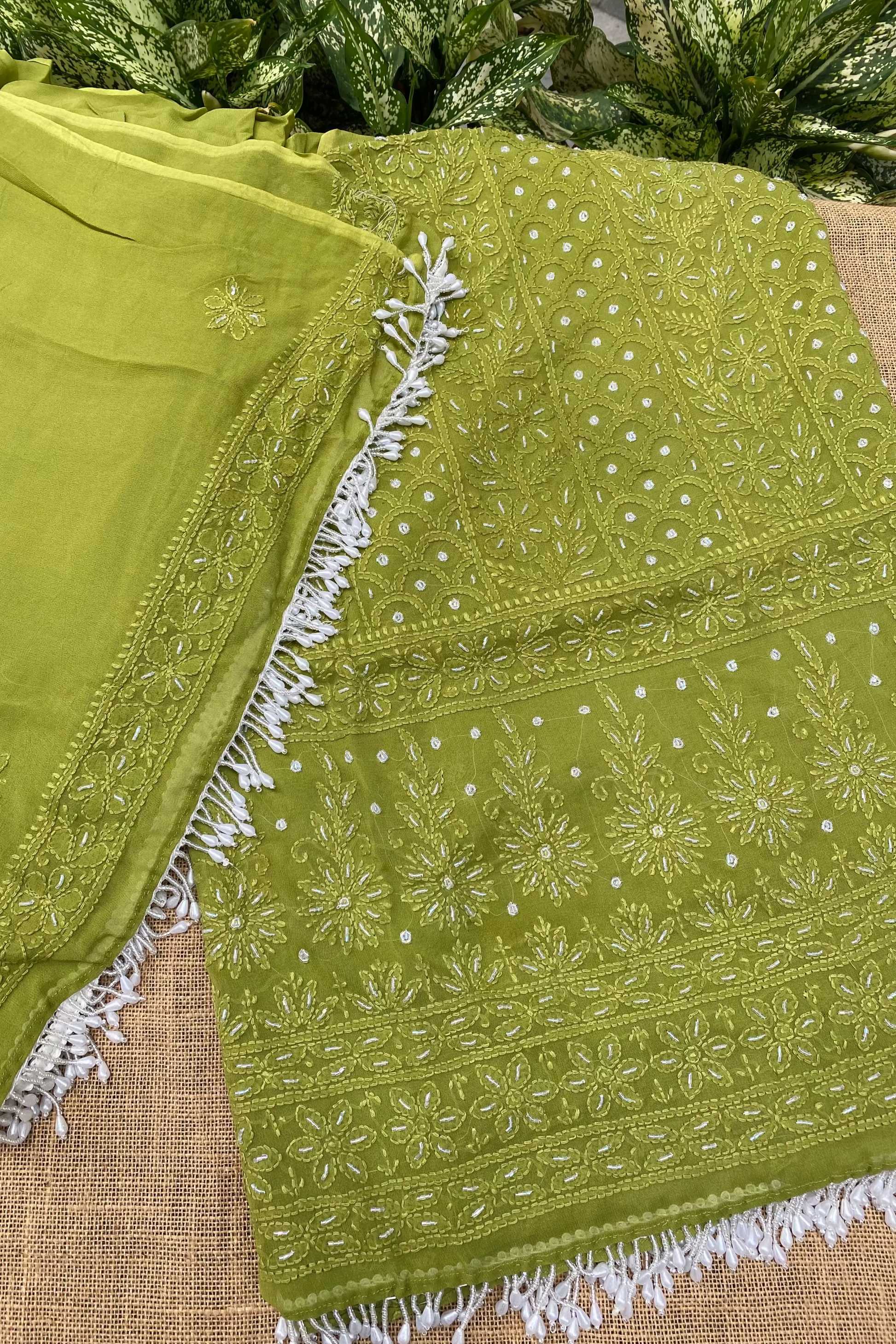An olive green embroidered fabric, likely a dupatta or scarf, featuring intricate white floral embroidery and white fringe trim, laid out on a burlap surface with green foliage in the background.