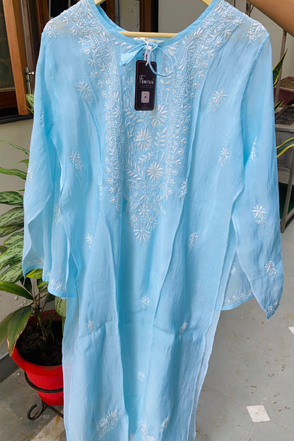 A light blue kaftan dress with white floral embroidery, hanging on a wooden hanger with a visible 'Tendai' brand tag, set against an indoor background with a potted plant.