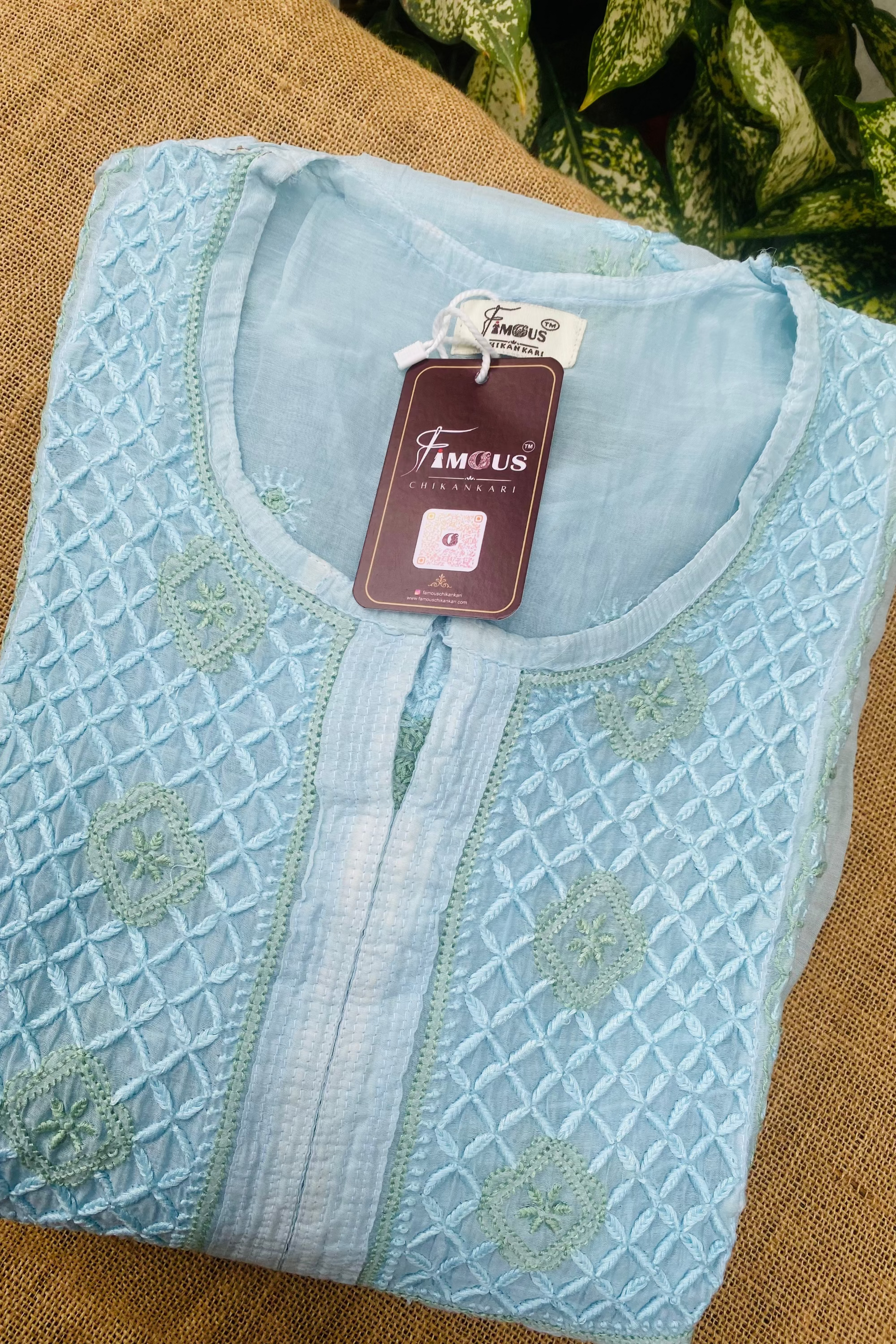 A folded light blue embroidered kurta with a geometric pattern and a split neckline, shown with a 'FAMOUS COLLECTION' tag on a burlap surface with green leaves in the background.