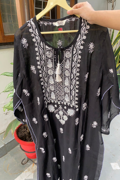 A black cotton kaftan tunic with white embroidery and tassel tie details, held up on a wooden hanger by a person's hand in an outdoor setting.