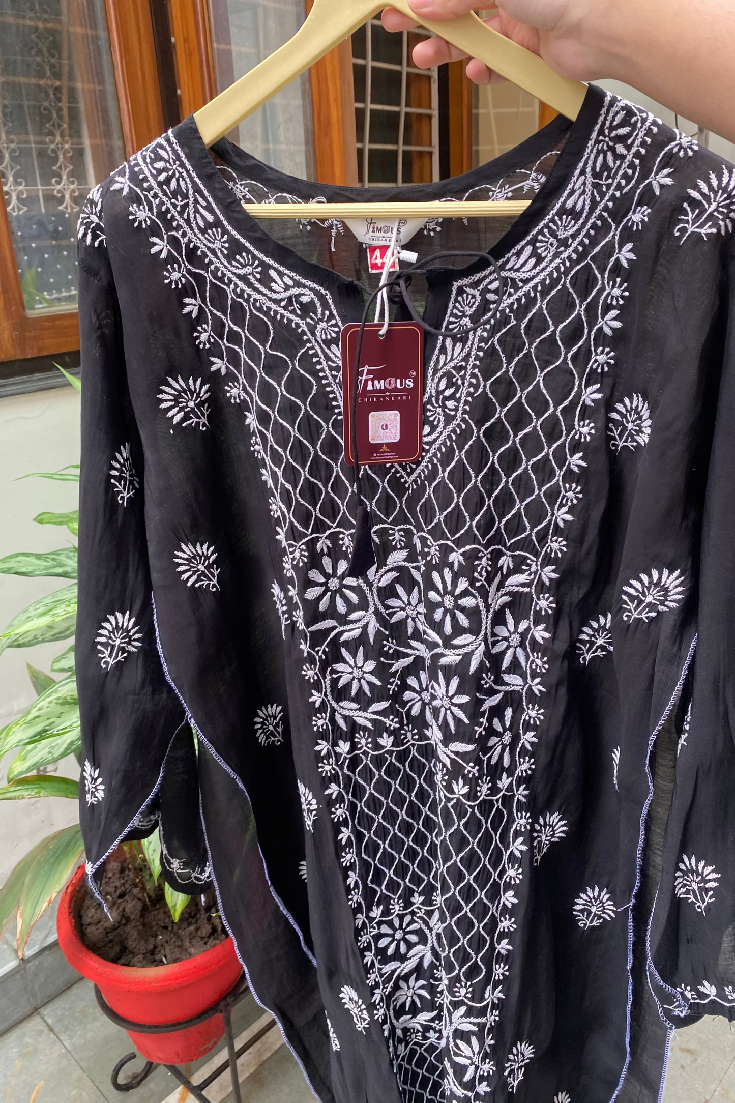A black long-sleeve tunic top with white floral and geometric embroidery, held on a wooden hanger by a hand. A red 'Femila' brand tag is attached to the neckline.