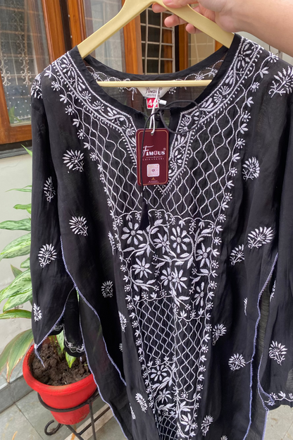 A black long-sleeve tunic top with white floral and geometric embroidery, held on a wooden hanger by a hand. A red 'Femila' brand tag is attached to the neckline.