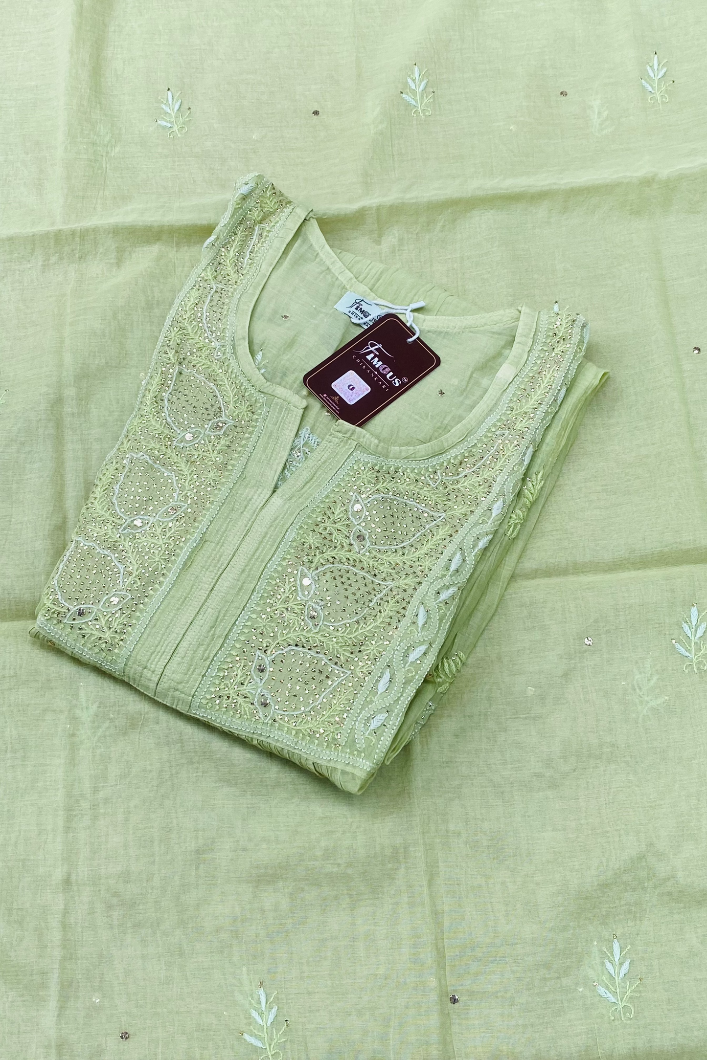 A folded light green embroidered kurta laid on matching fabric, featuring a round neckline with detailed floral embroidery and a small maroon brand tag.