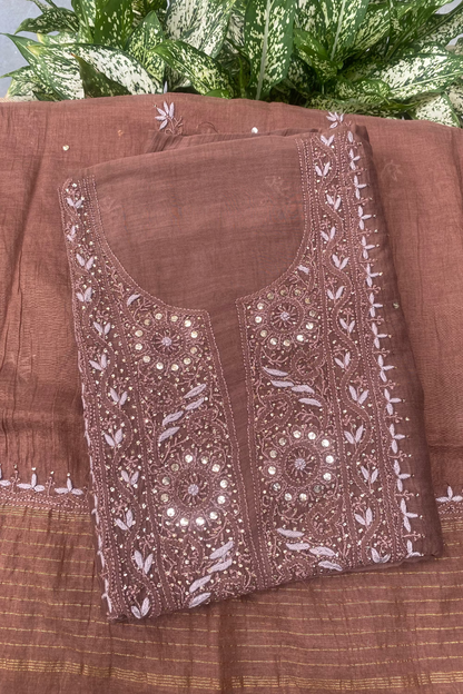 A folded brown salwar suit fabric set with detailed white and silver mirror work embroidery on the neckline, displayed on a matching striped brown dupatta with green foliage in the background.