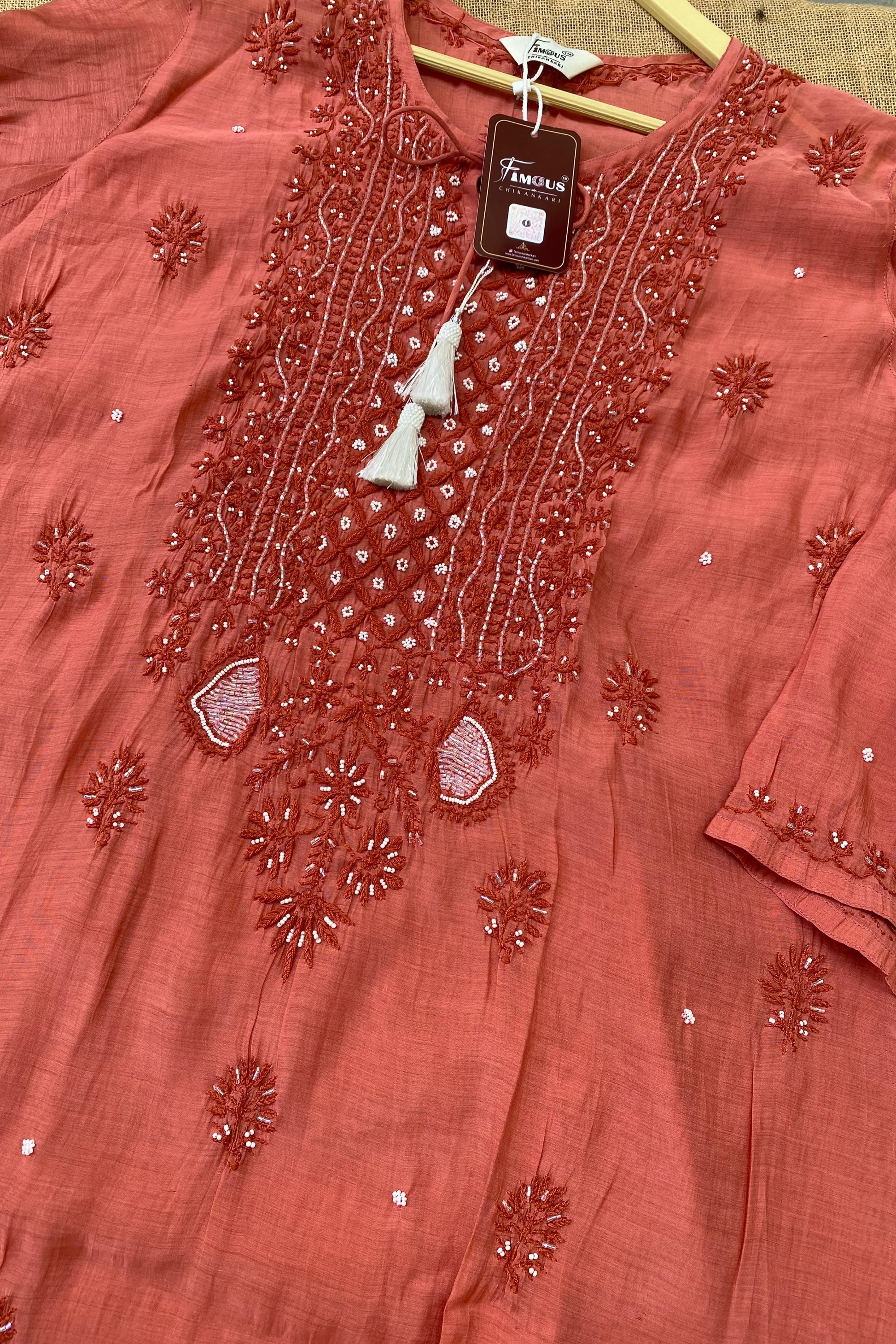 A terracotta pink embroidered kurta laid flat on a wooden hanger, featuring intricate floral embroidery, beadwork, and a white tassel detail on the neckline.