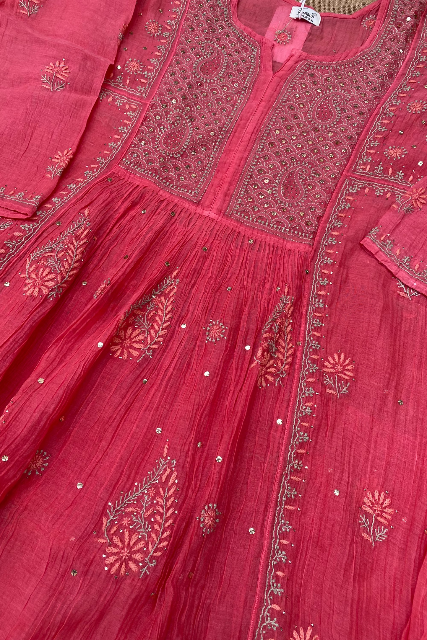 A close-up of a pink embroidered fabric garment, likely a kurta or dress, featuring detailed floral and paisley embroidery in matching pink and silver thread, with small mirror work accents.